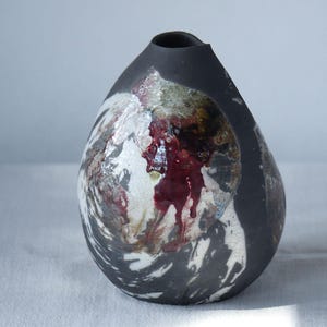 May include: A teardrop-shaped ceramic vase with a matte black exterior and a textured, crater-like opening. The surface features swirling white and grey patterns with a prominent, glossy, deep red glaze drip running down the center of the opening.