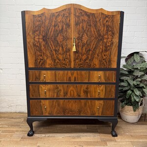 May include: A vintage wooden cabinet with a burled wood front. The cabinet has a dark grey frame, three drawers, and a curved top. Gold-toned hardware and black claw feet complete the piece. A classic storage solution, ideal for a living room or bedroom.
