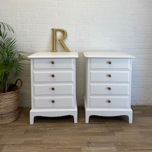 May include: Two white wooden chest of drawers with four drawers each. The drawers have gold knobs. The chests are on a wood floor.