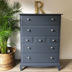 May include: A tall, grey chest of drawers with six drawers and silver knobs. The chest of drawers has a slightly curved base and sits on a wooden floor.