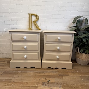 May include: A pair of cream-coloured bedside tables, each with three drawers and white, round knobs. The bases have a scalloped design. A gold letter 'R' is on the wall behind them. A potted plant in a woven basket is to the right.