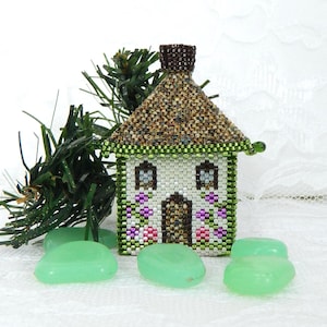 May include: A miniature beaded house with a brown thatched roof, white walls, and green trim. The house features small windows and a door, with purple and pink floral accents. Green glass stones and a sprig of evergreen are in the foreground.