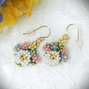 Può includere: Un paio di orecchini floreali fatti a mano. Ogni orecchino presenta un gruppo di piccole perline colorate bianche, rosa, lavanda, verdi e dorate, disposte a forma di fiore. Gli orecchini hanno ganci dorati.