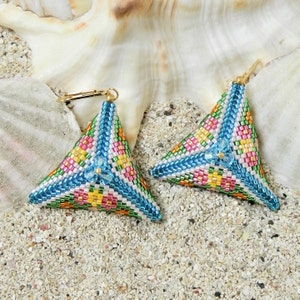 May include: A pair of triangle-shaped earrings made with colourful beads. The earrings are blue, green, pink, white, and yellow. The earrings are on a white sandy surface.