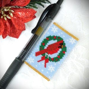 May include: A beaded Christmas ornament featuring a red cardinal bird inside a green wreath with red berries, set against a light blue background with white snowflakes. The ornament is rectangular with gold trim. A black pen rests beside it.