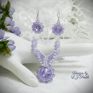 May include: A beaded jewelry set featuring a necklace and matching earrings. The set is primarily lavender with white accents. The necklace has a large, round pendant. The earrings are also round. The jewelry is displayed on a white surface.