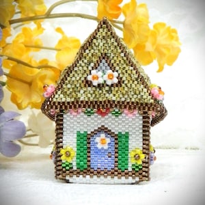 Może przedstawiać: Miniaturowy dom z koralików o szczegółowym wzorze. Dach jest brązowo-beżowy, ściany białe, a drzwi niebieskie. Zielone okiennice flankują drzwi, a małe kwiaty zdobią dom.