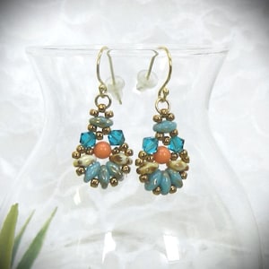 May include: A pair of drop earrings featuring a central round orange bead surrounded by small metallic gold seed beads, teal blue crystal bicones, and elongated light blue and cream-colored glass beads, suspended from gold-toned metal ear wires.