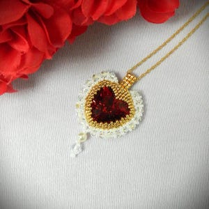 Könnte beinhalten: Halskette mit herzförmigem Anhänger. Der Anhänger hat ein rotes Herz, umgeben von goldenen Perlen und weißen Kristallen. Eine kleine Perle und ein Kristall hängen unten. Eine rote Blume befindet sich im Hintergrund.