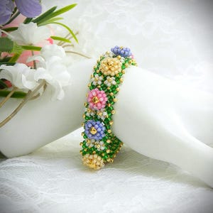 May include: A beaded bracelet featuring a floral design. The bracelet has a green base with colorful beaded flowers in pink, yellow, and blue. The flowers have gold centers. The bracelet is displayed on a white surface.