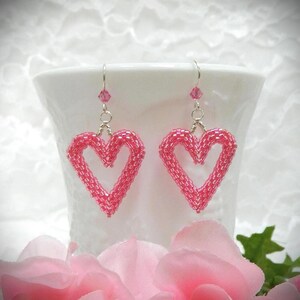 May include: A pair of dangle earrings featuring heart-shaped charms crafted from small, vibrant pink beads. Each earring hangs from a silver-toned metal hook and includes a single, small, faceted pink crystal bead positioned above the heart.