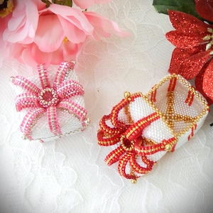 May include: Two beaded gift boxes. One is square with a pink bow, the other is rectangular with a red and gold bow. Both boxes are white with beaded accents. Pink and red flowers are in the background.