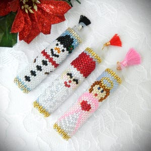 May include: Three decorative bottle stoppers with cross-stitched Christmas designs: a snowman, Santa Claus, and an angel. Each stopper has a gold-colored trim and a tassel. The snowman has a black top hat and a red scarf. The angel wears a halo.