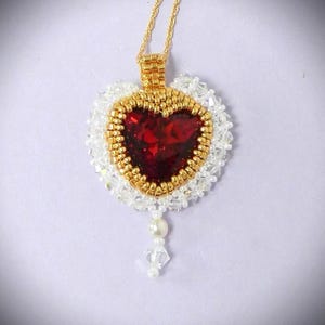 May include: A heart-shaped pendant necklace with a gold chain. The pendant features a large, faceted red gemstone heart, surrounded by gold beads and white seed beads. A small pearl and crystal dangle from the bottom.