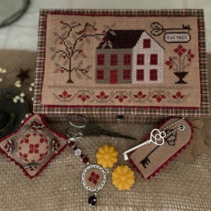 Può includere: Un kit di ricamo a punto croce con una casa rossa e bianca con un tetto marrone, un albero con foglie rosse e un vaso di fiori con fiori rossi. Il kit include una scatola di legno, un salvan ago, un portachiavi e un ciondolo con perline.