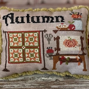 Può includere: Un cuscino beige con un bordo a frange verdi presenta un disegno a punto croce che rappresenta una scena autunnale. Il cuscino riporta la scritta "Autumn" in lettere nere e include un blocco di trapunta, una macchina da cucire e una torta.