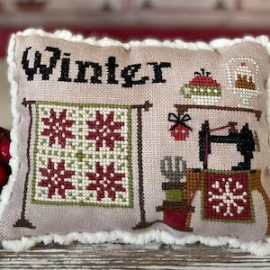 Può includere: Un cuscino beige con un bordo bianco e soffice presenta un disegno a punto croce di una scena invernale. Il disegno include un blocco di trapunta con fiocchi di neve rossi e verdi, una macchina da cucire e la parola "Winter" in lettere nere.