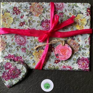 Può includere: Un regalo avvolto in carta floreale con rose rosa, legato con un nastro rosa brillante. Sono visibili anche un sottobicchiere a forma di cuore e un'etichetta rotonda con un motivo a rosa.