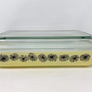 Peut inclure: Un plat de cuisson rectangulaire en verre jaune avec un couvercle en verre transparent. L'extérieur du plat présente un motif répétitif de fleurs de marguerite dessinées au trait noir sur les côtés. Le plat possède des poignées intégrées.