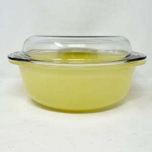 May include: A vintage yellow Pyrex casserole dish with a clear glass lid. The dish has a rounded shape with a slight lip and a clear glass lid. The dish is a pale yellow colour and appears to be made of glass. The lid is clear and fits snugly on top.