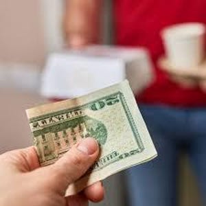 May include: A hand holding a twenty-dollar bill, featuring Andrew Jackson's portrait. The bill is creased, with green and tan hues. In the background, a person is holding food and drinks.