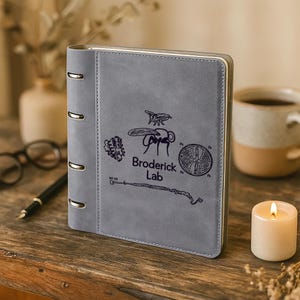 May include: A gray, leather-bound ring binder notebook featuring a dark, printed graphic of a fly, a circular pattern, and the text Broderick Lab. The notebook has four silver-colored metal rings along the left spine and a smooth, matte finish.