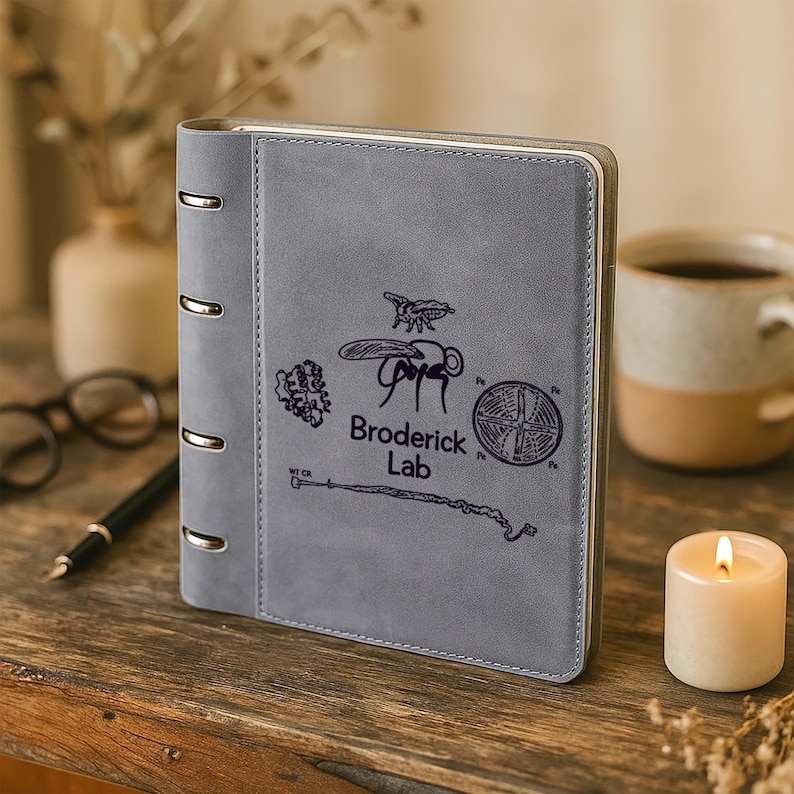 May include: A gray, leather-bound ring binder notebook featuring a dark, printed graphic of a fly, a circular pattern, and the text Broderick Lab. The notebook has four silver-colored metal rings along the left spine and a smooth, matte finish.