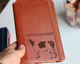 May include: A brown leather-bound journal with the words "Cattle Records" embossed on the front cover. A black and white illustration of a cow is also engraved on the cover.