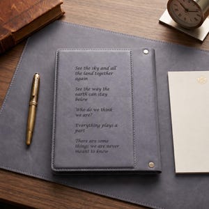 May include: A grey leather-bound journal featuring a vertical stitched panel and two silver-toned metal rivets. The cover is engraved with the text: See the sky and all the land together again. See the way the earth can stay below. Who do we think we are? Everything plays a part. There are some things we are never meant to know.