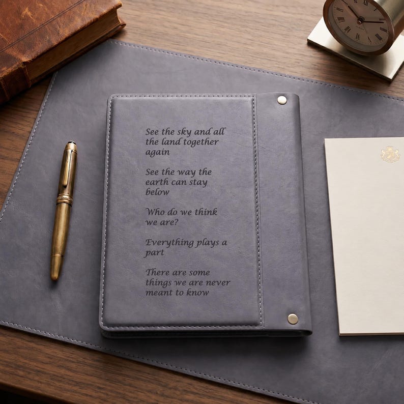 May include: A grey leather-bound journal featuring a vertical stitched panel and two silver-toned metal rivets. The cover is engraved with the text: See the sky and all the land together again. See the way the earth can stay below. Who do we think we are? Everything plays a part. There are some things we are never meant to know.