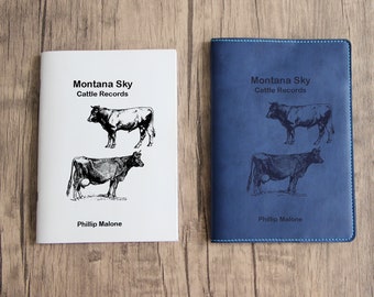 May include: Two different versions of a book titled "Montana Sky Cattle Records" by Phillip Malone. The book on the left is a white paperback with black text and illustrations of cows. The book on the right is a blue hardcover with black text and illustrations of cows.
