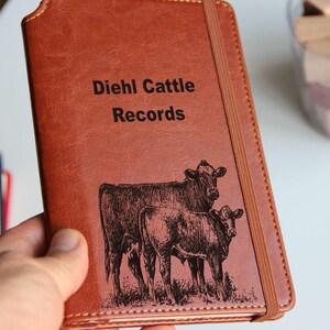 May include: A brown leather-bound journal with the text "Diehl Cattle Records" embossed on the front cover. The cover also features an engraved image of two cows, one standing behind the other.