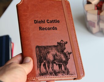 May include: A brown leather-bound journal with the text "Diehl Cattle Records" embossed on the front cover. The cover also features an engraved image of two cows, one standing behind the other.