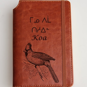 以下が含まれることがあります： 茶色の革製ジャーナル。カーディナルバードのイラストと「Koa」の文字が特徴です。ジャーナルはゴムバンドで閉じられ、縁はステッチされています。表紙には、未知の言語のテキストも含まれています。