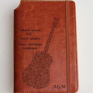 May include: A brown leather journal with a guitar made of musical notes engraved on the front. The quote "Where words fail, music speaks. Hans Christian Andersen" is also engraved on the front. The journal has a brown elastic closure and the initials "AGM" are engraved on the bottom right corner.