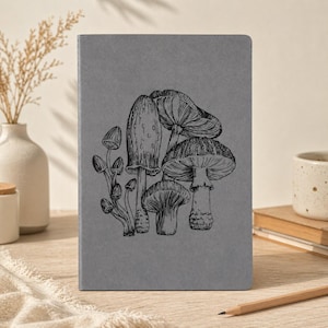 May include: A grey notebook featuring a black and white illustration of different mushrooms. The journal is closed and has a textured cover. A pencil, a vase with dried plants, and a mug are in the background.