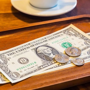 May include: Two one-dollar bills and several coins sit on a wooden tray. The bills feature a portrait of George Washington. A white saucer and the edge of a coffee cup are visible in the background.