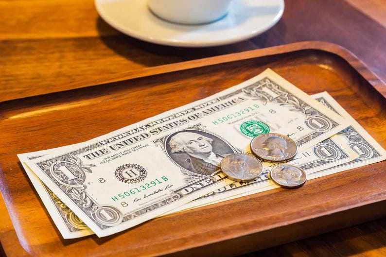May include: Two one-dollar bills and several coins sit on a wooden tray. The bills feature a portrait of George Washington. A white saucer and the edge of a coffee cup are visible in the background.