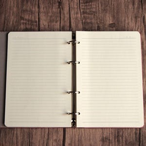May include: An open ring binder notebook with a grey cover and cream-colored lined paper. The interior features a silver-toned metal four-ring mechanism centered between two pages of horizontal ruled lines, providing a flat surface for writing.