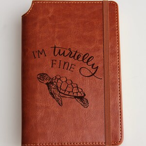May include: A brown leather journal with an elastic closure. The front cover features an engraved image of a sea turtle and the text "I'm turtlely fine".