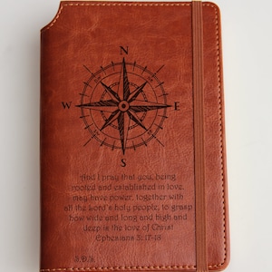 May include: Brown leather journal with a compass and a quote from Ephesians 3:17-18. The quote reads: "And I pray that you, being rooted and established in love, may have power, together with all the Lord's holy people, to grasp how wide and long and high and deep is the love of Christ Ephesians 3:17-18"