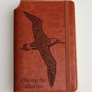 May include: A brown leather journal with an engraved image of an albatross in flight. The journal has a brown elastic closure and the text "Chasing the albatross" is engraved below the image.