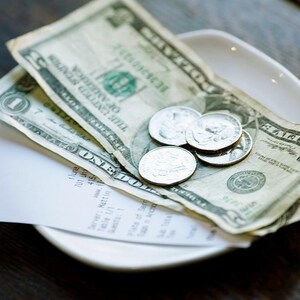 May include: A white ceramic plate holds a tip of paper currency and coins. The bills are a mix of one and five-dollar denominations. Several silver coins are also visible. A white receipt is partially visible.