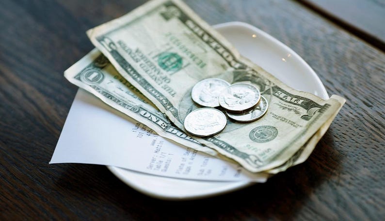 May include: A white ceramic plate holds a tip of paper currency and coins. The bills are a mix of one and five-dollar denominations. Several silver coins are also visible. A white receipt is partially visible.