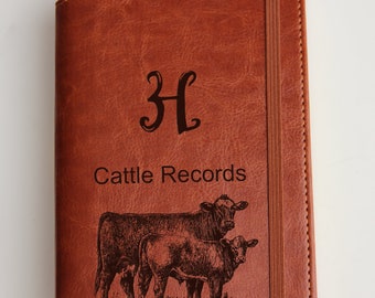 May include: Brown leather journal with a personalized monogram "H" and the text "Cattle Records" engraved on the front. The journal features an engraved image of two cows and a calf.