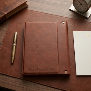May include: A brown leather-like notebook featuring a vertical stitched panel on the right side with two silver-toned metal rivets. The cover is personalized with the engraved text Nichole A. Broderick, PhD in a cursive script font.