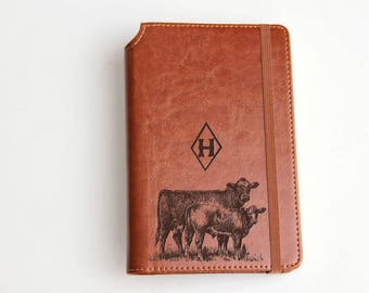 May include: A brown leather journal with an embossed design of a cow and calf. The journal has a diamond-shaped emblem with the letter "H" inside. A brown elastic band secures the journal.