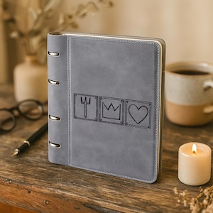 May include: A gray, matte-finish ring binder notebook featuring a vertical spine with four silver metal rings. The front cover displays three square-bordered engravings: a trident, a crown, and a heart, each framed with small star and moon symbols.