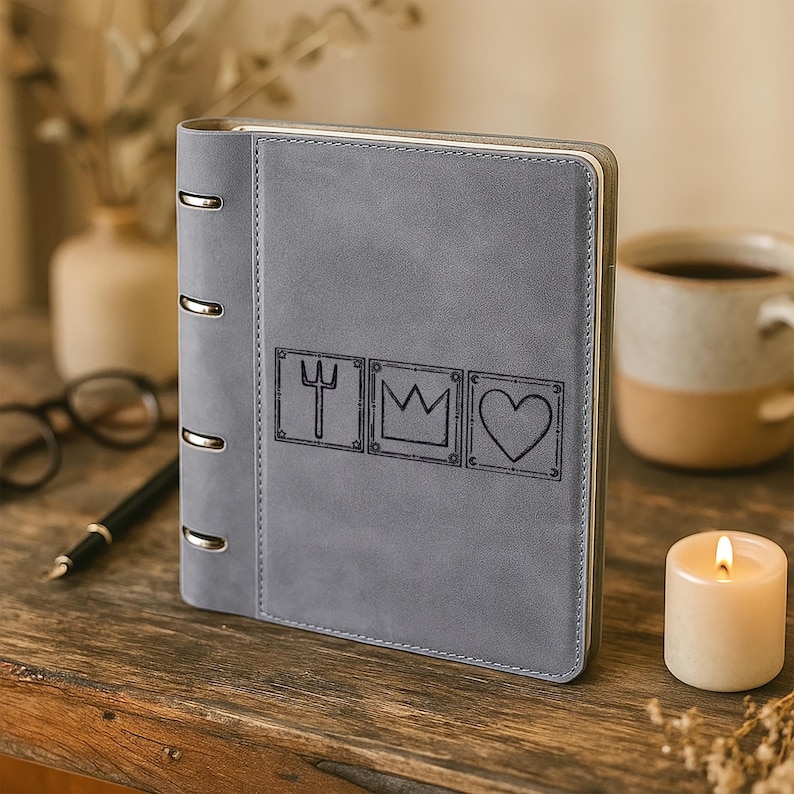 May include: A gray, matte-finish ring binder notebook featuring a vertical spine with four silver metal rings. The front cover displays three square-bordered engravings: a trident, a crown, and a heart, each framed with small star and moon symbols.