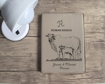 Puede incluir: Un diario de cuero marrón con un diseño grabado en negro de una vaca y un ternero. El diario tiene el texto "Roman Ranch" y "Javier & Manuel Roman" grabado en la parte delantera.